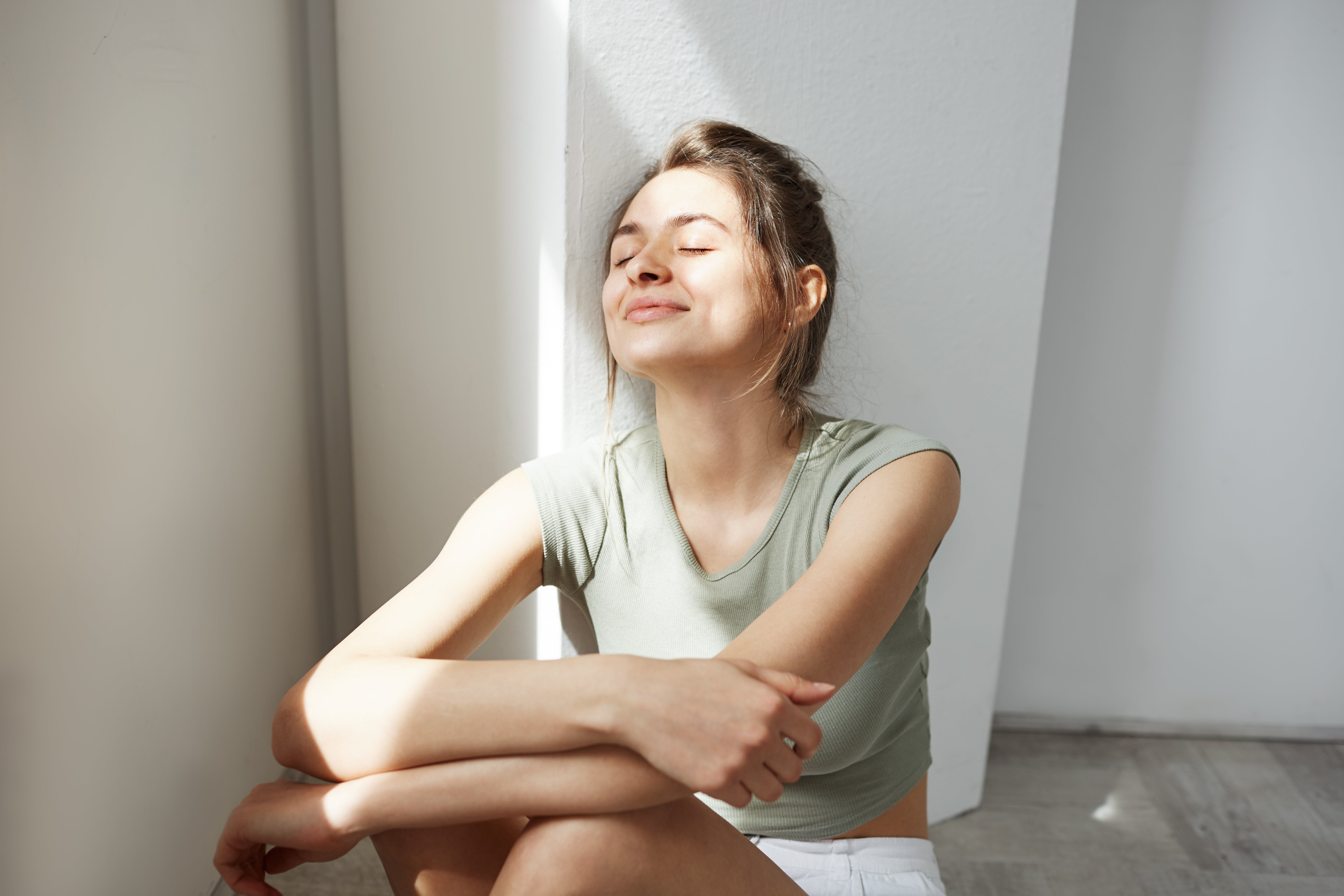 Young woman smiling with her eyes closed, relaxing under sun rays against a wall